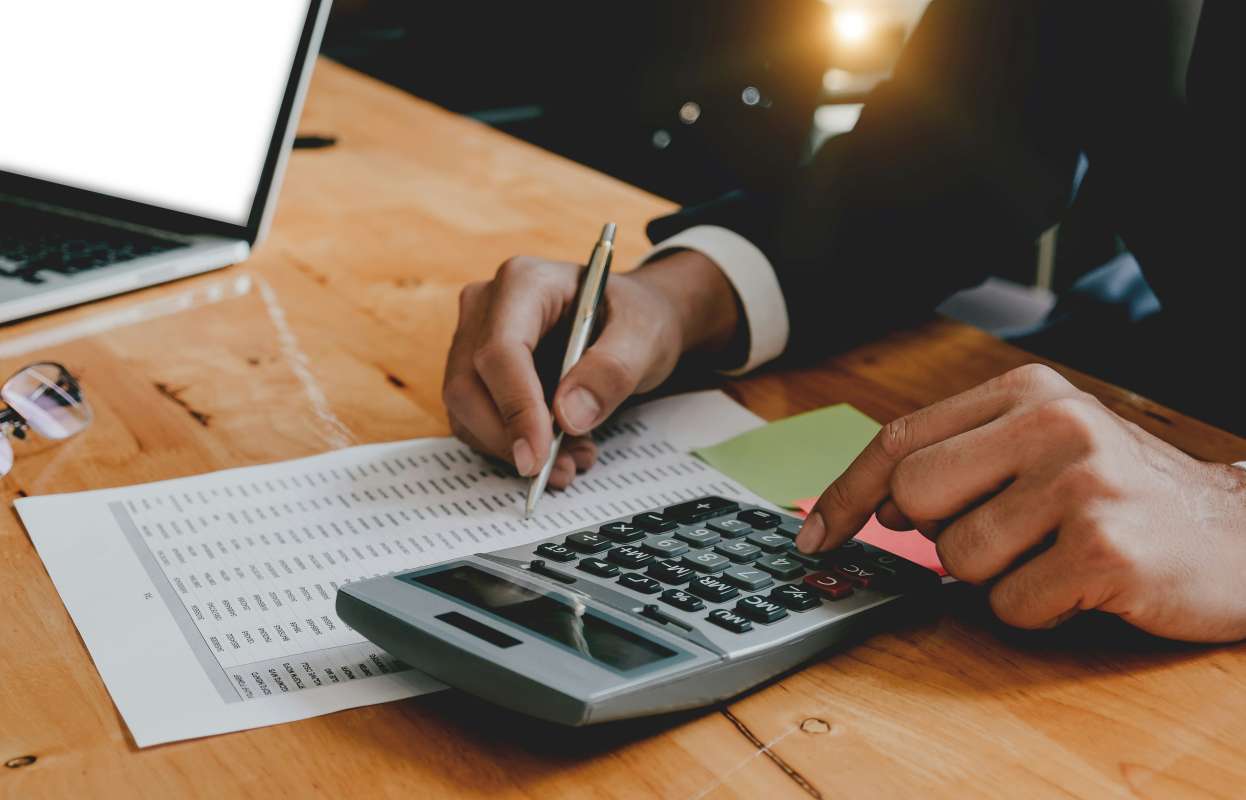 Pessoa em um escritório, sentada em uma mesa de madeira, focada em cálculos. Com a mão direita, a pessoa está usando uma calculadora de mesa grande, enquanto a mão esquerda segura uma caneta sobre uma planilha ou documento cheio de números e dados.